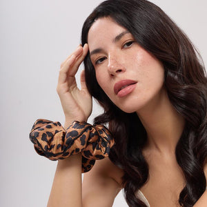A woman with long, voluminous hair wears the Hotel Paradiso - Wild Thing Padded Scrunchie by us.drowsysleepco on her wrist. With natural makeup and visible freckles, she poses confidently, gazing at the camera.