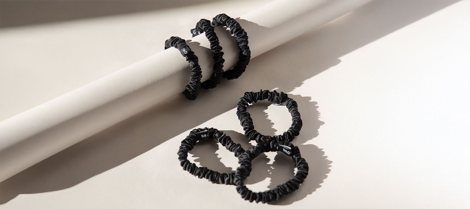 Four black scrunchies are displayed, with two wrapped around a white cylindrical rod and two placed on a light beige surface, casting soft shadows in natural light.