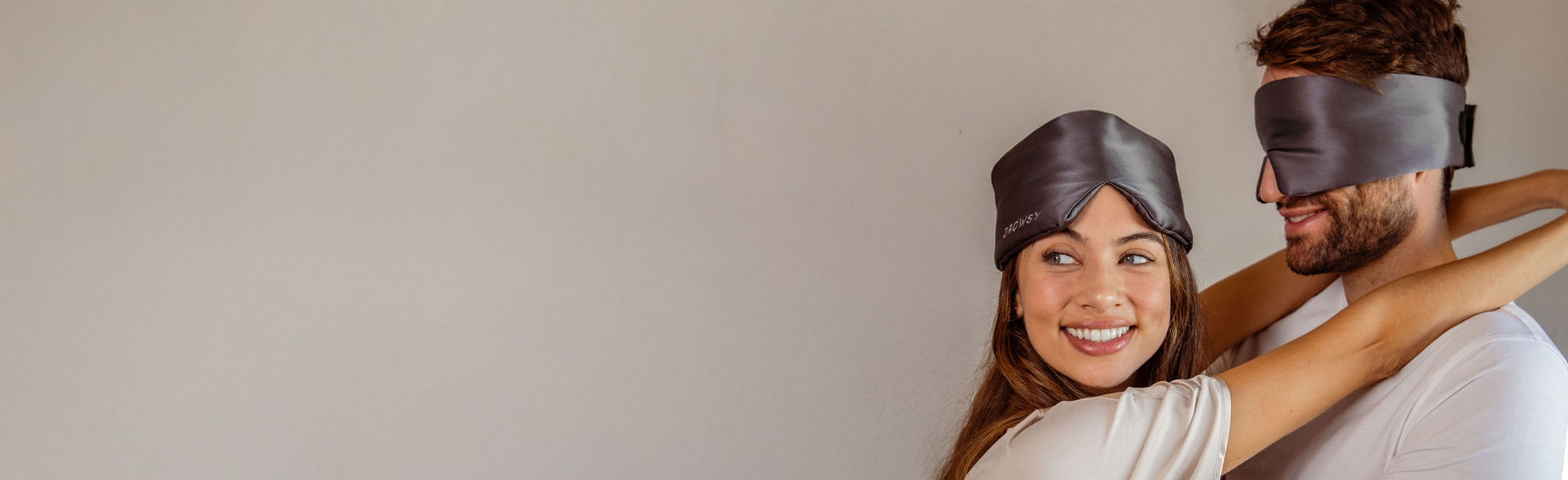 A smiling woman and a man, both wearing gray sleep masks, embrace each other while standing against a plain light-colored background.
