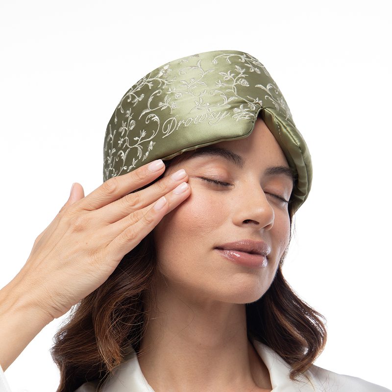 A person with eyes closed wears a Pride & Prejudice - Sage + English Rose Eyelash Protecting Mask, a sage green mulberry silk sleep mask featuring white floral embroidery and the word Drowsy embroidered on the front.