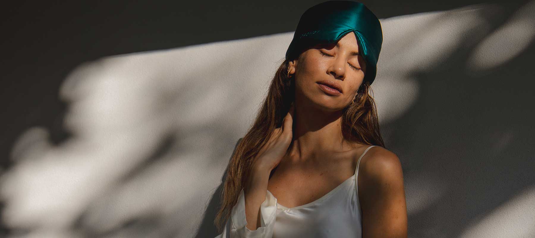 A woman wearing a dark green satin sleep mask and a white silk camisole stands against a sunlit wall with shadows, gently touching her neck and closing her eyes peacefully.