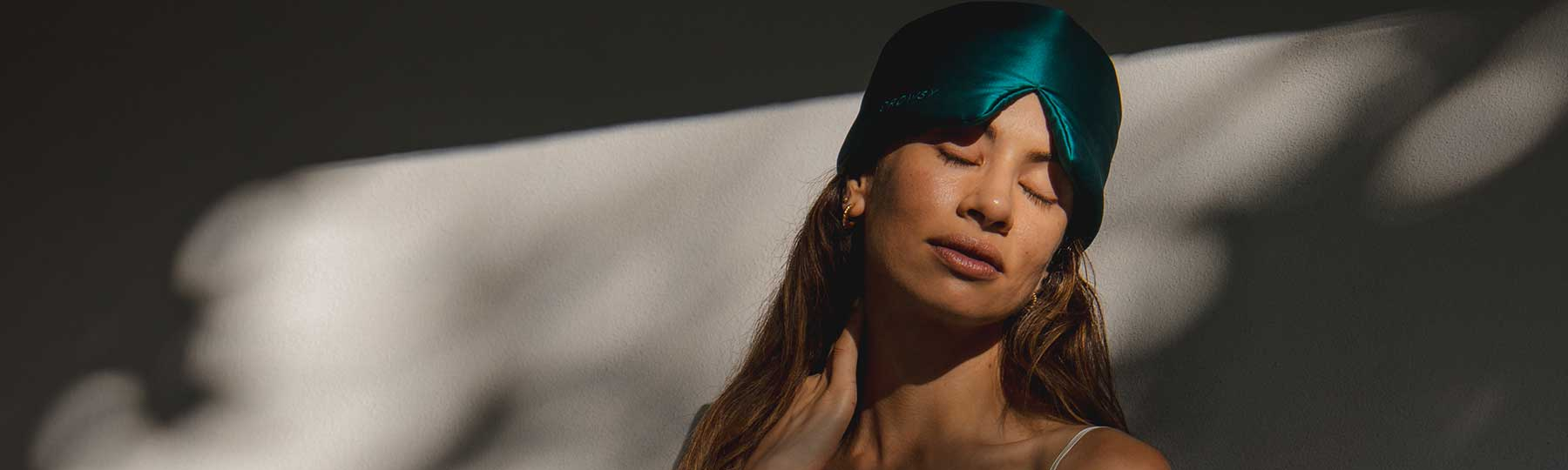 A woman with long hair and a teal satin sleep cap closes her eyes peacefully, standing in soft sunlight with shadows in the background.