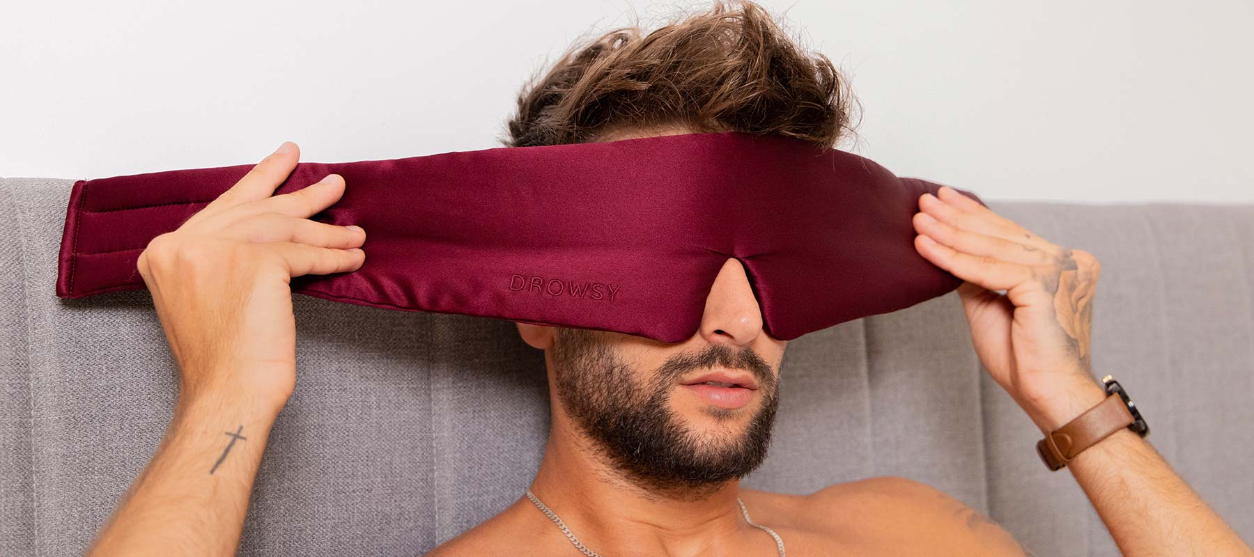 A man sits on a gray headboard bed, holding a burgundy eye mask over his eyes with both hands. He has short brown hair, facial hair, and is wearing a wristwatch and a bracelet.
