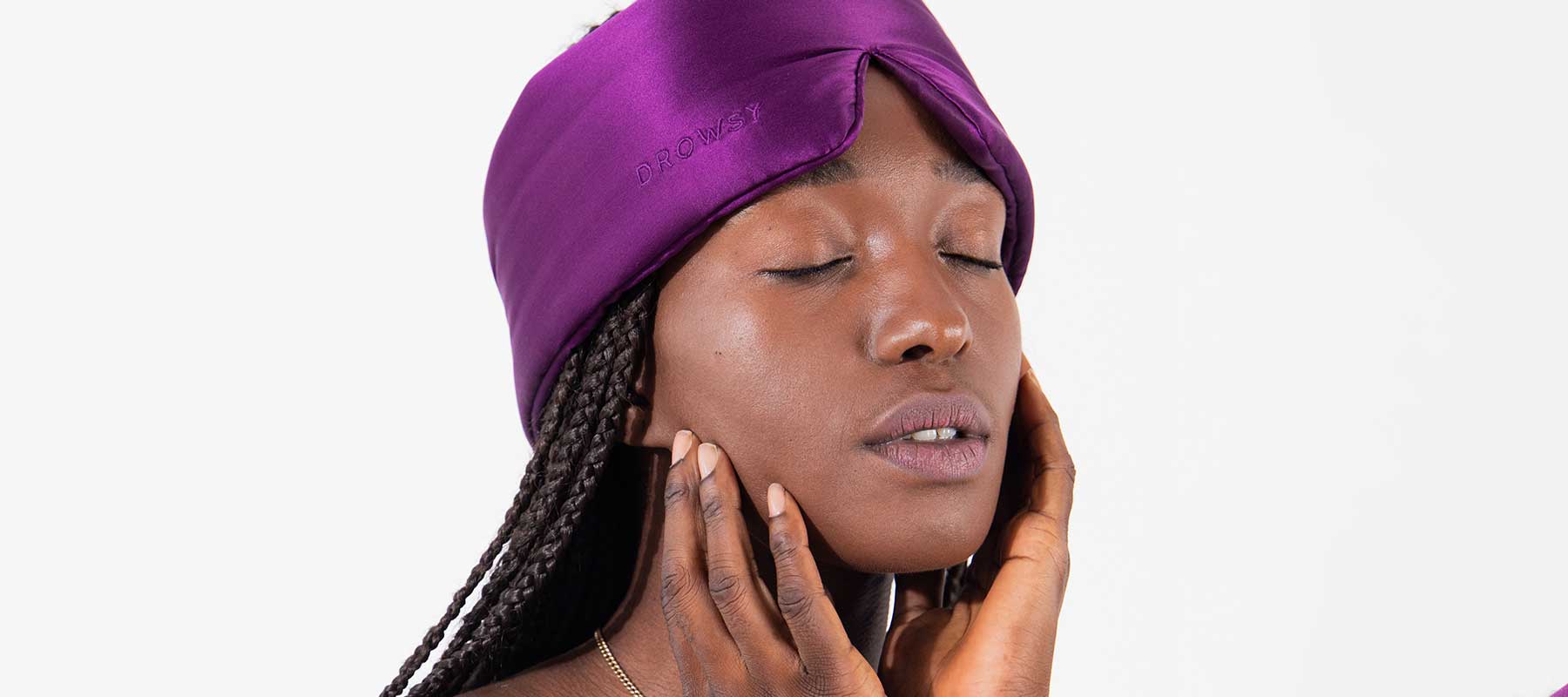 A woman with braided hair and closed eyes gently touches her face while wearing a purple silk sleep mask on her forehead against a plain white background.