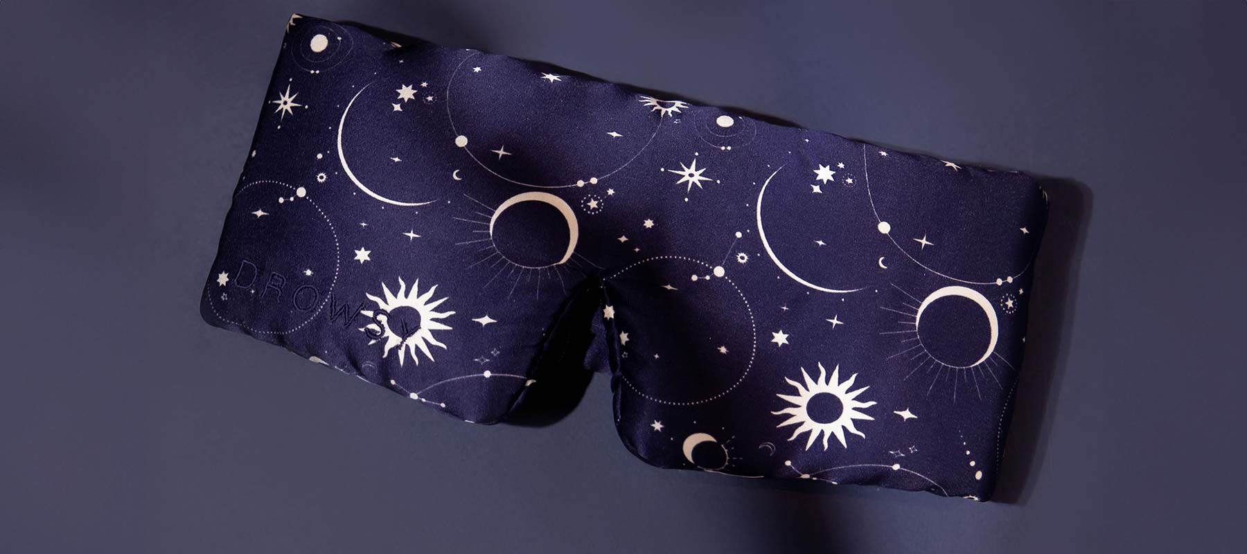 A dark blue eye mask with white celestial designs featuring moons, stars, and suns, displayed on a matching dark background.