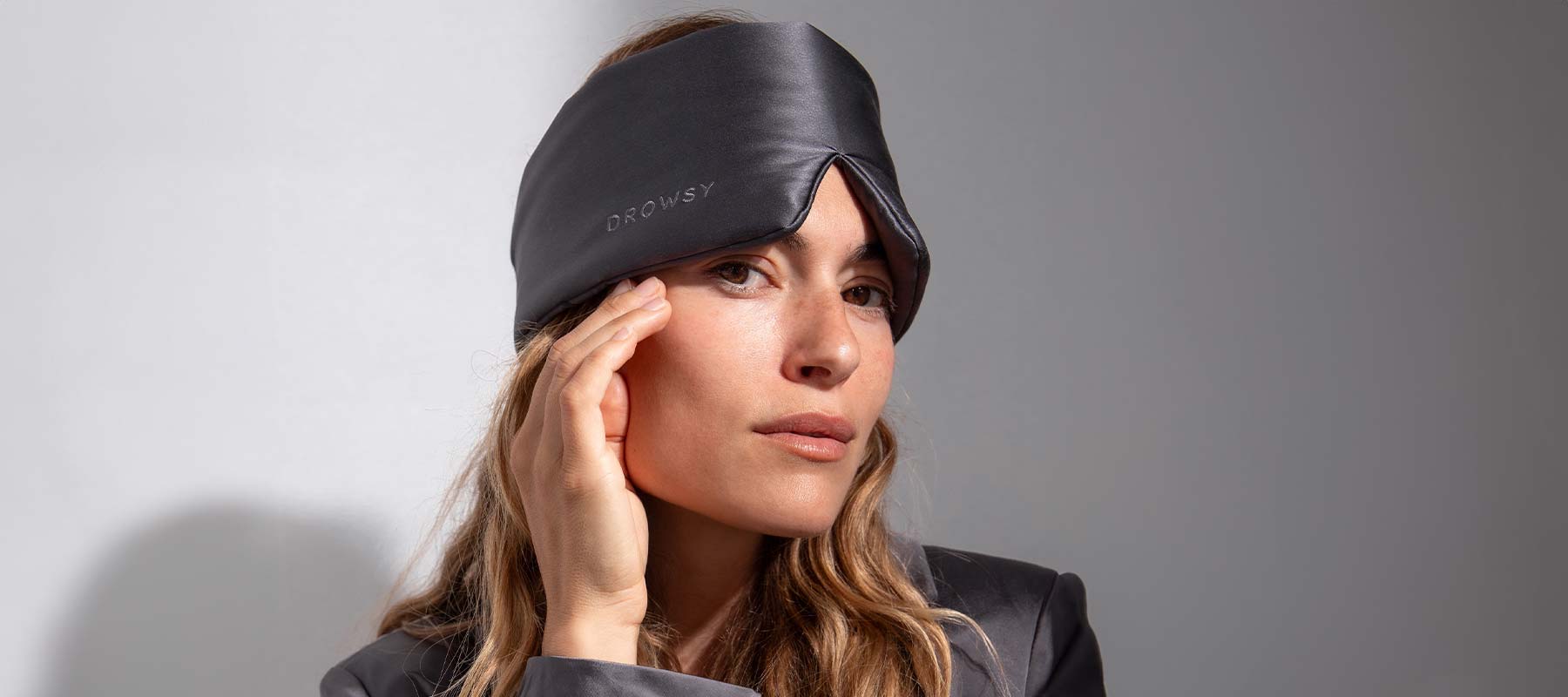 A woman wearing a black silk sleep mask and dark pajamas touches her face and gazes forward, standing against a plain light background.