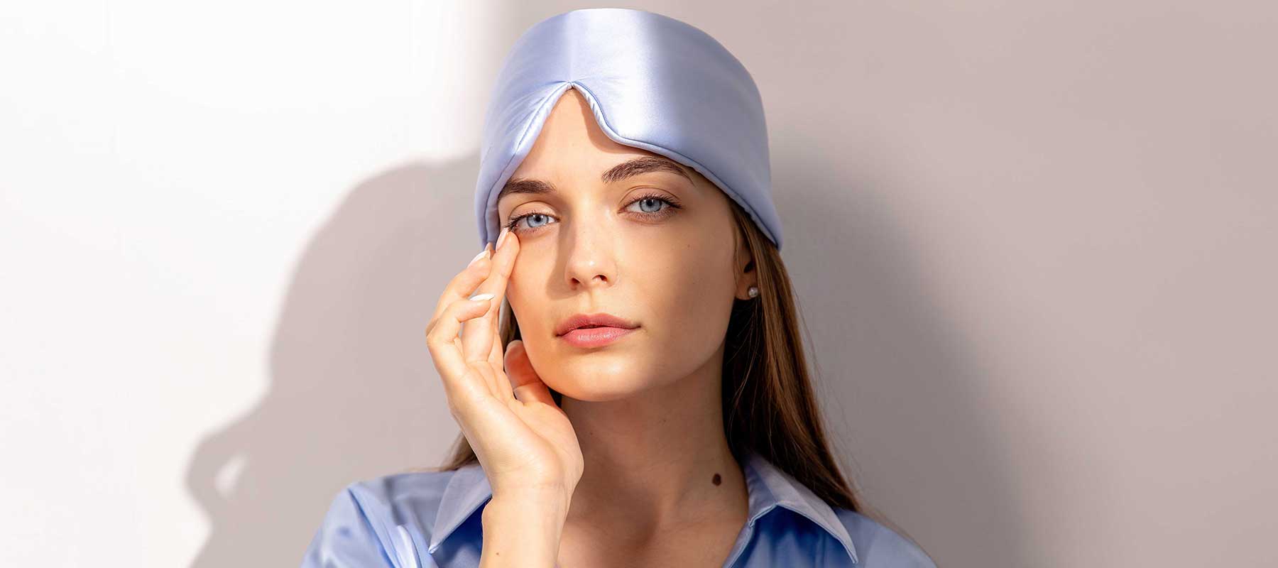 A woman with long hair wears a shiny light blue sleep mask on her head and a matching shirt. She gently touches her face and looks directly at the camera against a light background.
