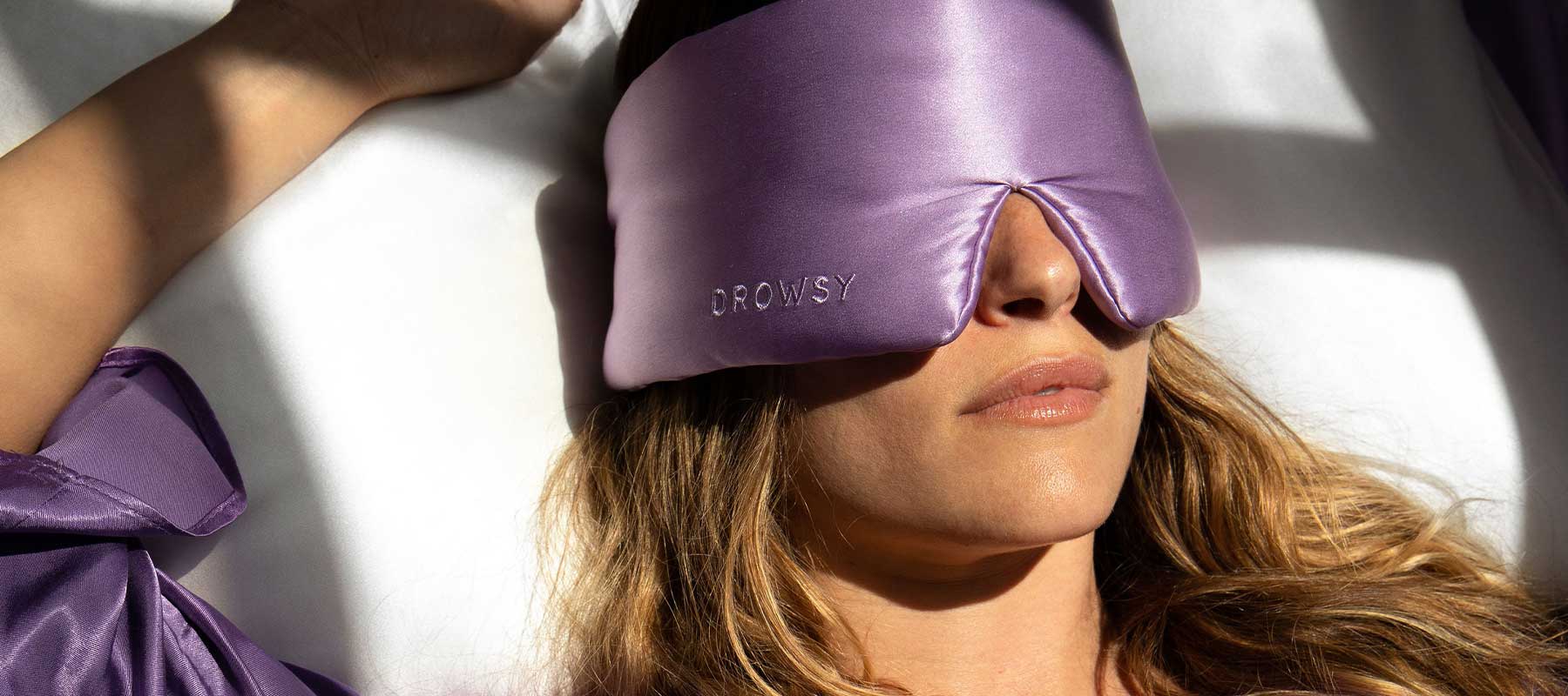 A person wearing a purple satin sleep mask with the word Drowsy on it lies in bed, dressed in matching purple sleepwear, with sunlight casting shadows across their face.