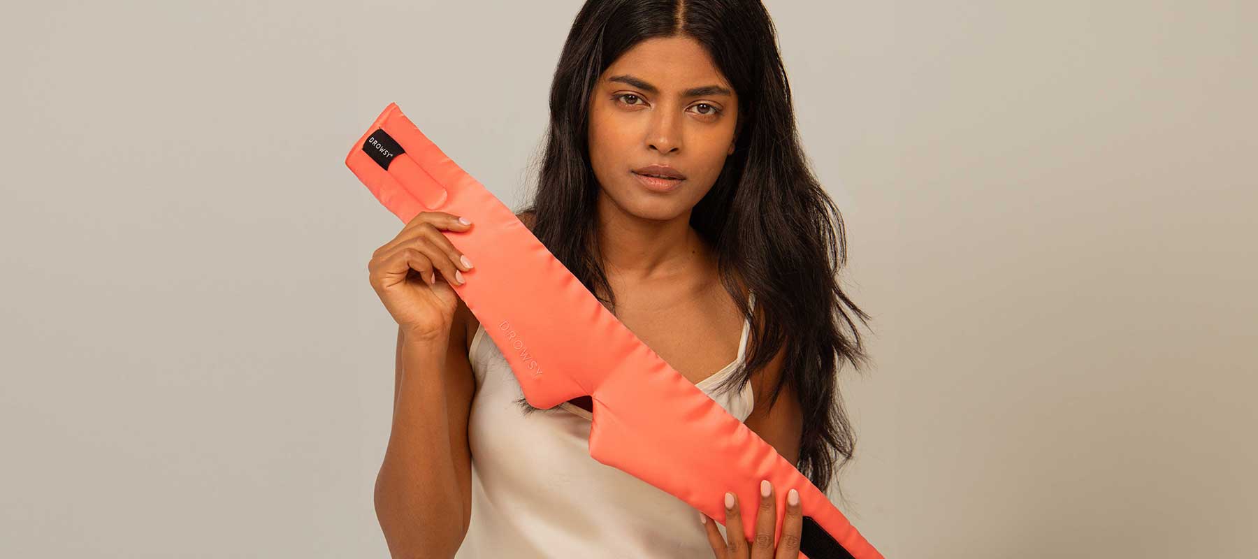 A woman with long dark hair and a neutral expression holds a coral-colored, padded wrap with a Velcro strap against her chest, standing in front of a plain light background.