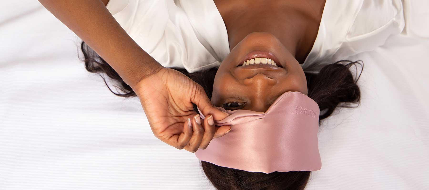 A woman lying on her back in bed smiles while adjusting a pink silk sleep mask on her forehead, wearing a white top and resting on white sheets.