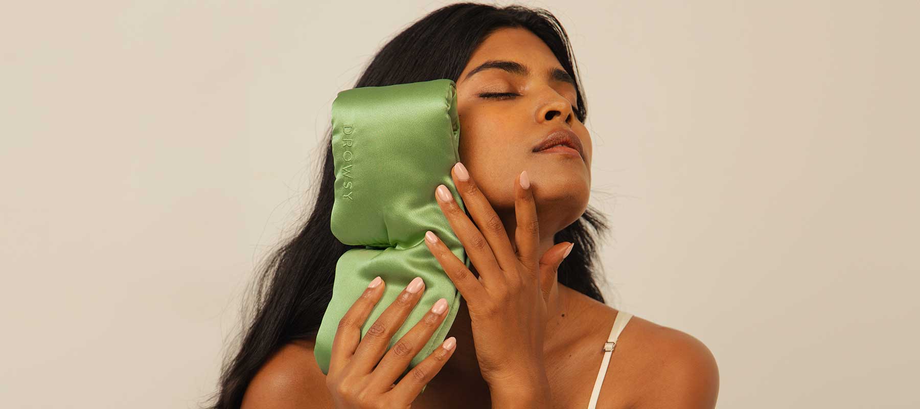 A woman with long dark hair presses a green satin ice pack to the side of her face, eyes closed, wearing a beige tank top against a neutral background.