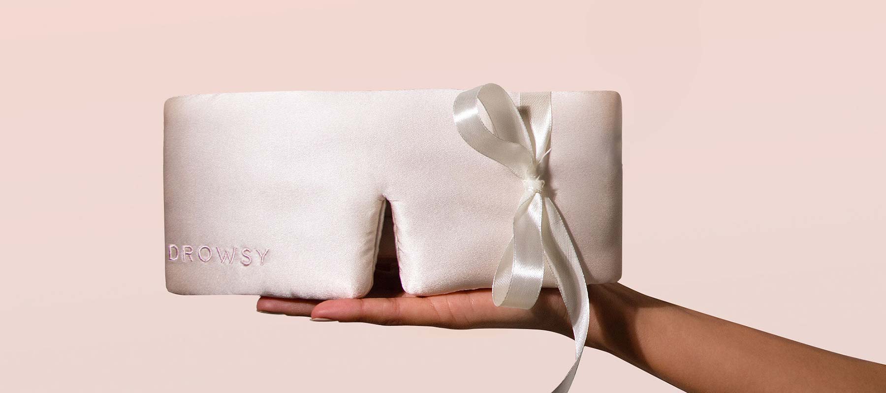 A hand holds a pale pink satin sleep mask with the word DROWSY embroidered on it and a white ribbon tied in a bow, against a soft pink background.