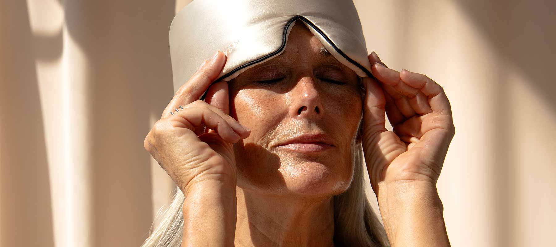 An older woman with long gray hair adjusts a satin sleep mask on her forehead, eyes closed, standing in soft natural light with a calm, relaxed expression.