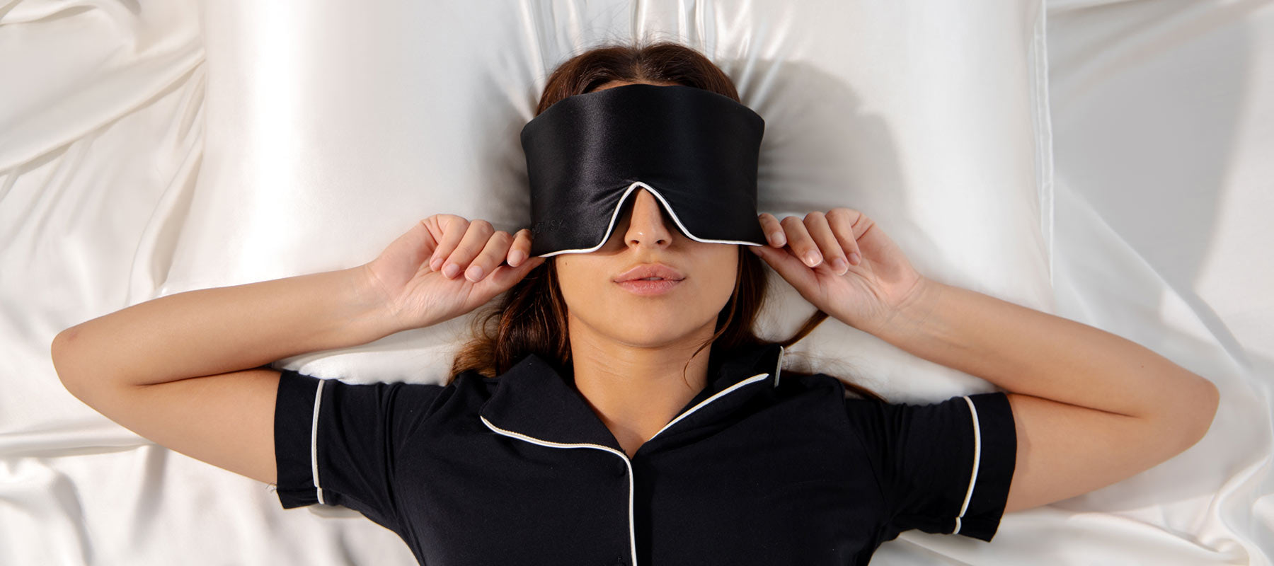 A woman lying on a white bed wears a black sleep mask and dark pajamas with white trim, holding the sides of the mask as she rests on a satin pillow.