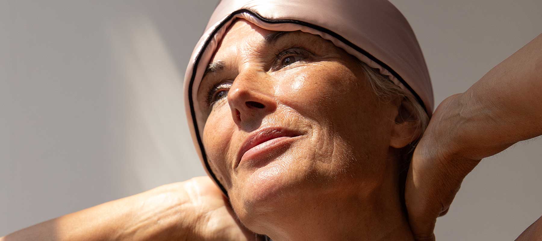 Close-up of an older woman with light skin, wearing a pink satin sleep mask pushed up on her forehead, looking upward with a relaxed expression and hands behind her neck in soft, natural light.