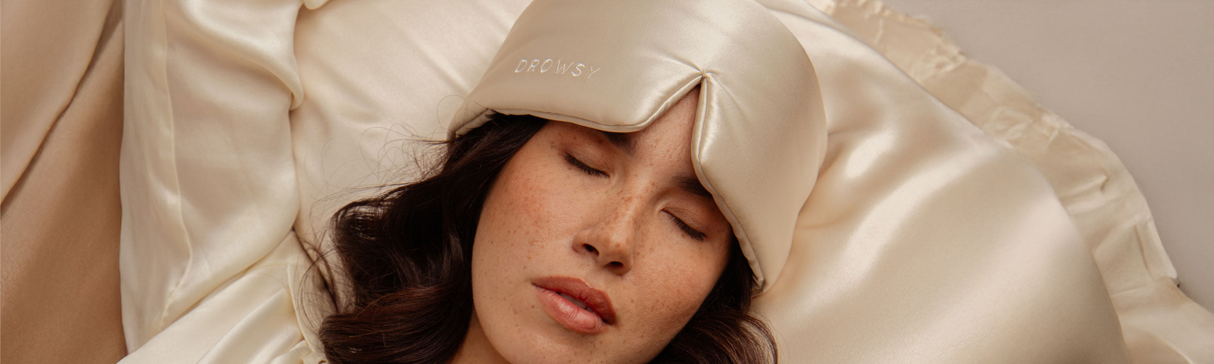 A woman with closed eyes rests on cream-colored satin bedding, wearing a matching satin sleep mask with DROWSY embroidered on it.