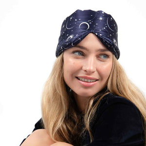 A smiling woman with long blonde hair wears us.drowsysleepcos Cosmic Energy dark blue turban-style headband, featuring a celestial blackout design with moons and stars, set against a white background.