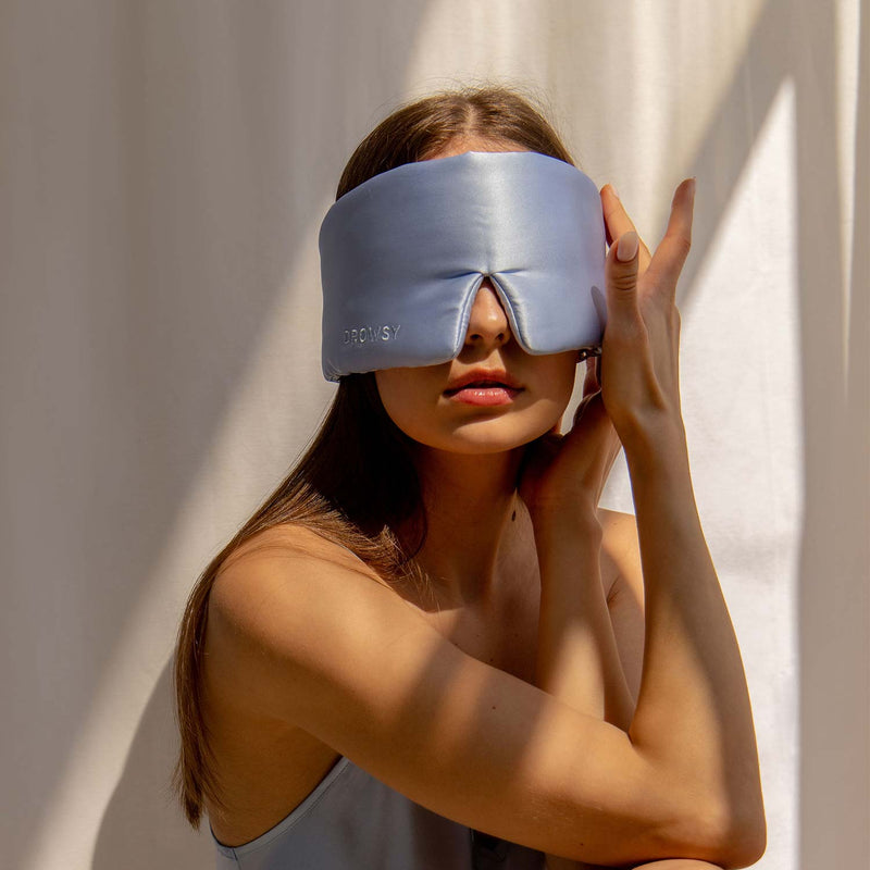 Model wearing Drowsy baby blue coloured silk sleep mask on a white background in the sunlight