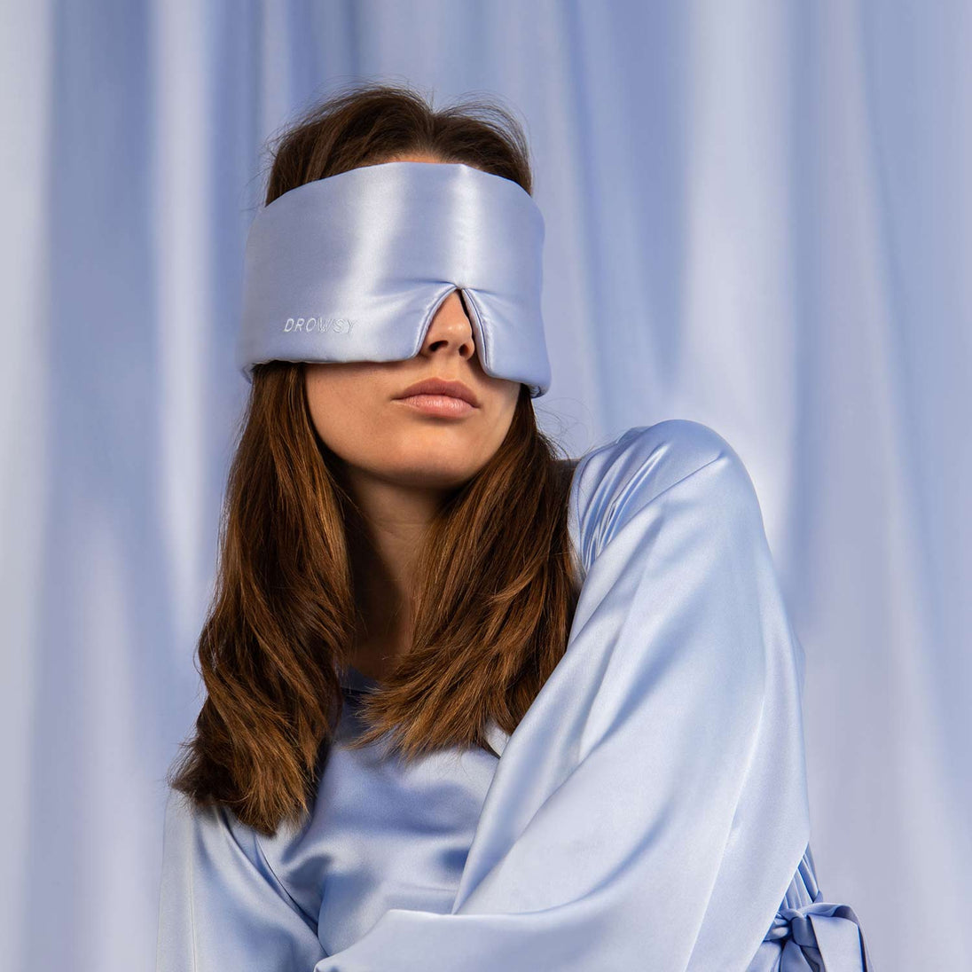 Model wearing blue silk silk sleep mask with a silk curtain backdrop
