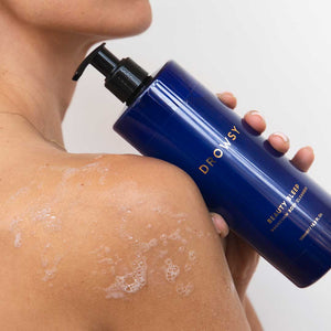 A person with damp, soapy skin holds a large blue pump bottle of us.drowsysleepco Beauty Sleep Body Cleanser against their shoulder—a calming magnesium night-ritual body wash—set on a white background.