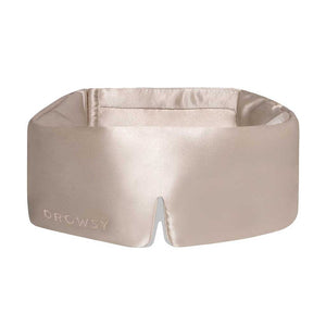 The Dusty Gold eye mask by us.drowsysleepco is a beige silk mask with DROWSY in small letters on the left, featuring a wide, cushioned blackout design and a small notch at the bottom center.