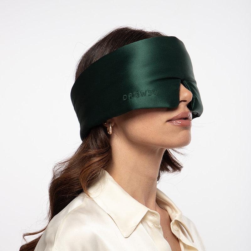 A woman with wavy brown hair is dressed in a cream-colored blouse, wearing the drowsysleepco Ivy Green padded silk sleep mask over her eyes against a plain background.