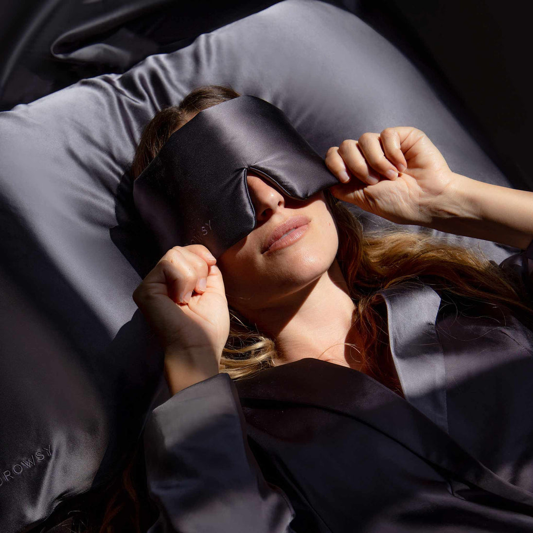 Model lying on grey silk sheets pulling a grey Drowsy silk sleep mask over her eyes