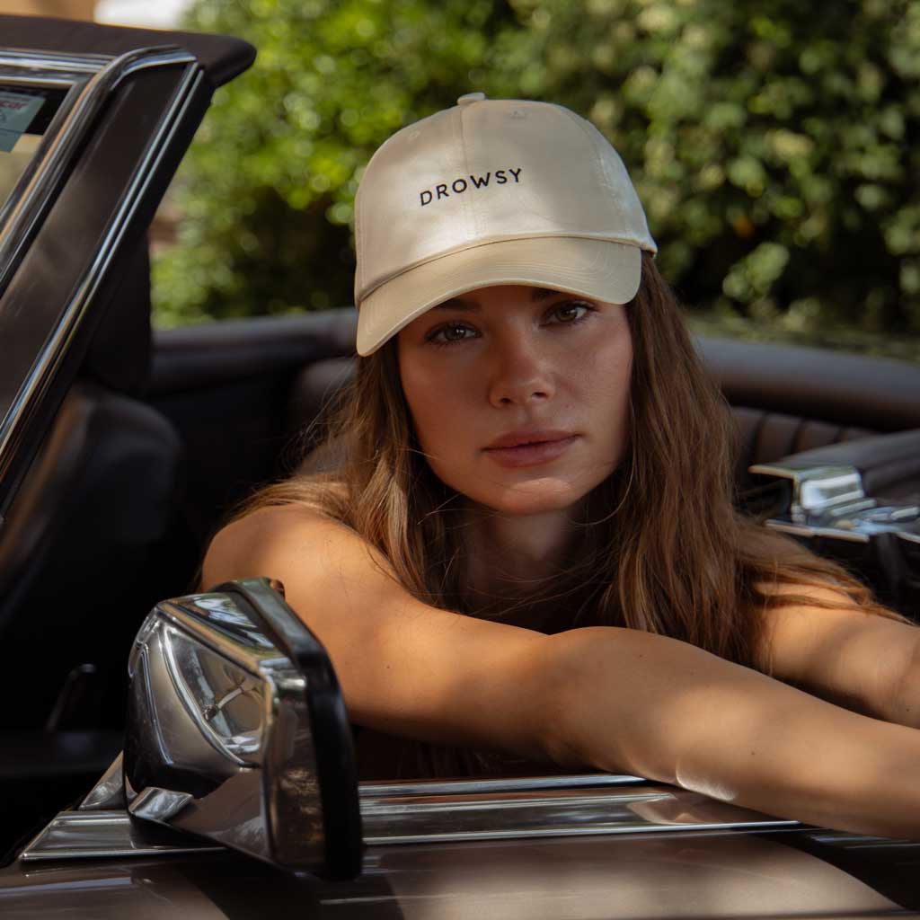 A woman wearing the Drowsy Silk Cap in Dusty Gold by Drowsy relaxes in a convertible, resting her arms on the door and gazing at the camera. The cap shields her face while greenery blurs in the background.