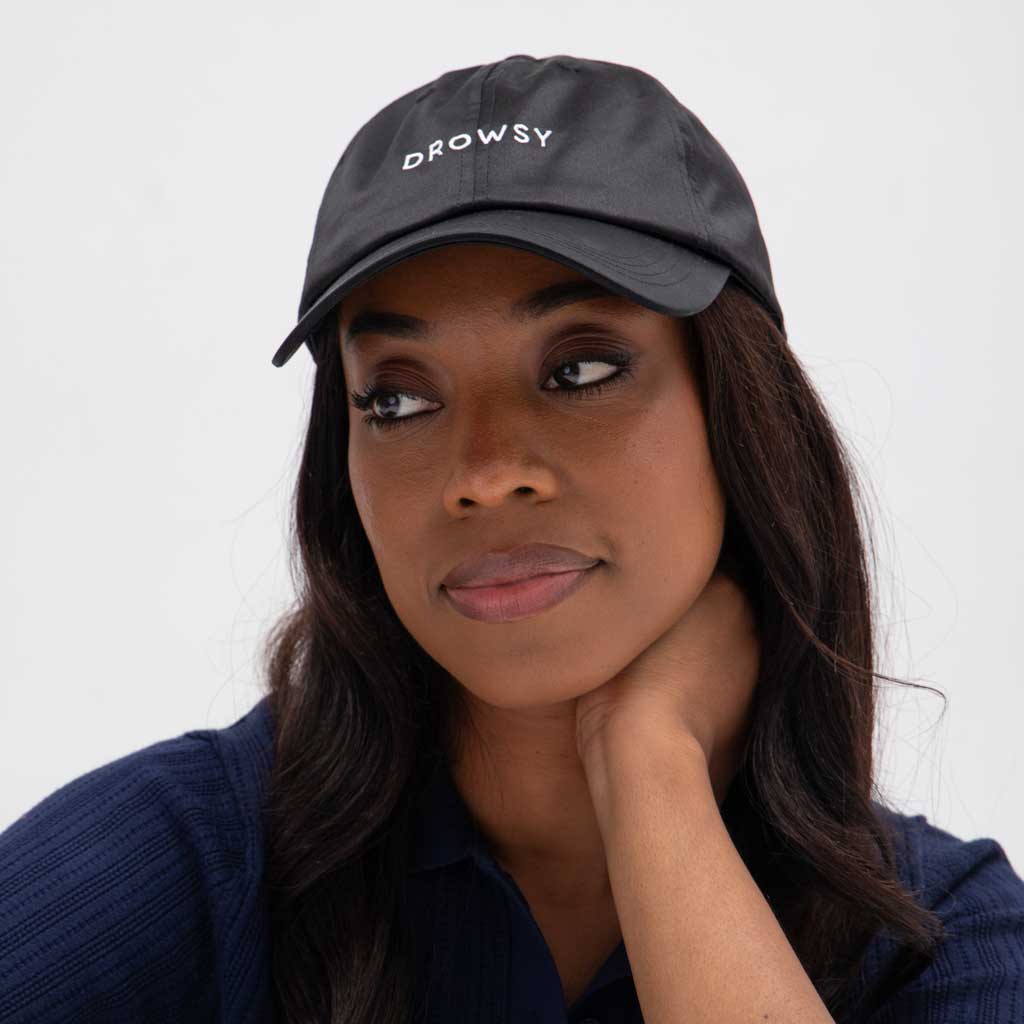 A woman with long dark hair wears the Drowsy Silk Cap - Moonlight Shadow by Drowsy, featuring “DROWSY” in white letters. She looks to the side with her hand on her neck, dressed in a dark shirt and a neutral expression.
