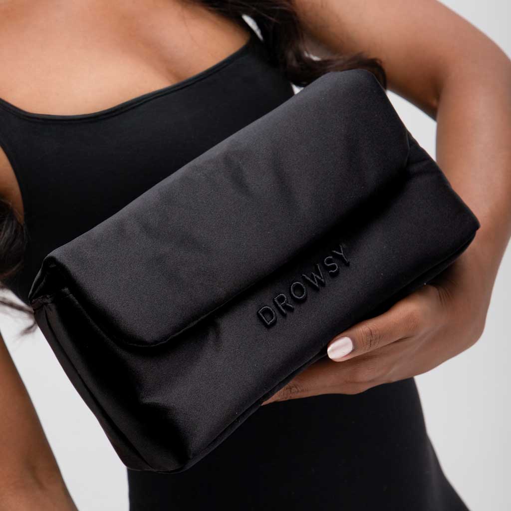 A person in a black sleeveless top holds the Drowsy Silk Necessaire - Black Jade, a soft and quilted skincare pouch by Drowsy, embossed with DROWSY on the front—ideal for organizing your beauty essentials.
