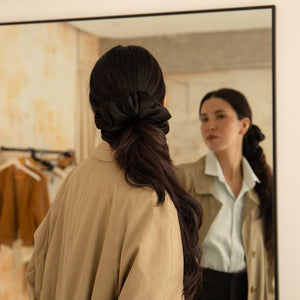 A woman with long dark hair tied back with the drowsysleepco Black Jade - Padded Scrunchie gazes at her reflection in a mirror, wearing a beige coat in a room filled with hanging clothes.
