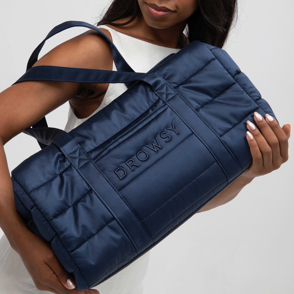 A woman in a white sleeveless outfit holds the Drowsy Roadtrip Bundle in Midnight Blue + Damask Rose, a quilted bag with a soft, padded look, two shoulder straps, and the Drowsy brand embossed on the side.