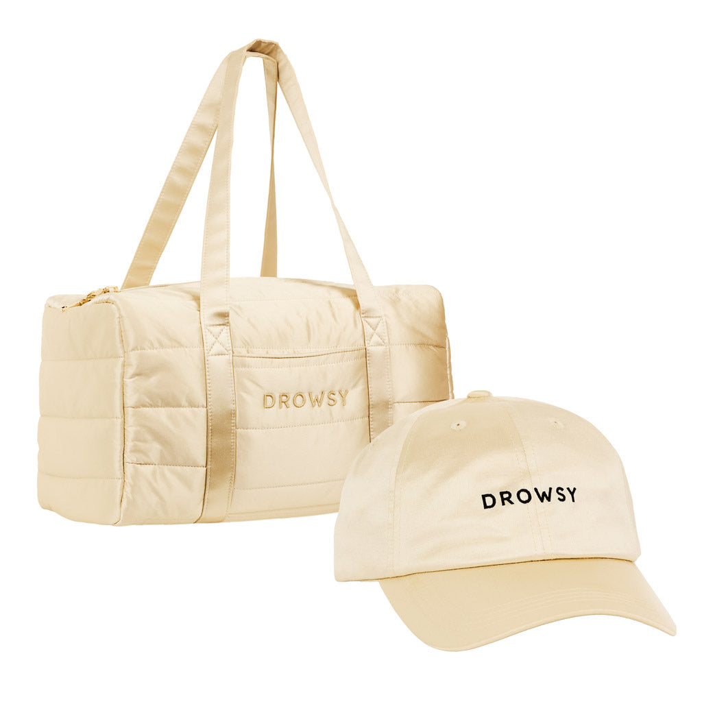 The Drowsy Roadtrip bundle in Dusty Gold includes a beige duffel bag and matching baseball cap, both featuring “DROWSY” embroidered in black on the front, set against a white background.