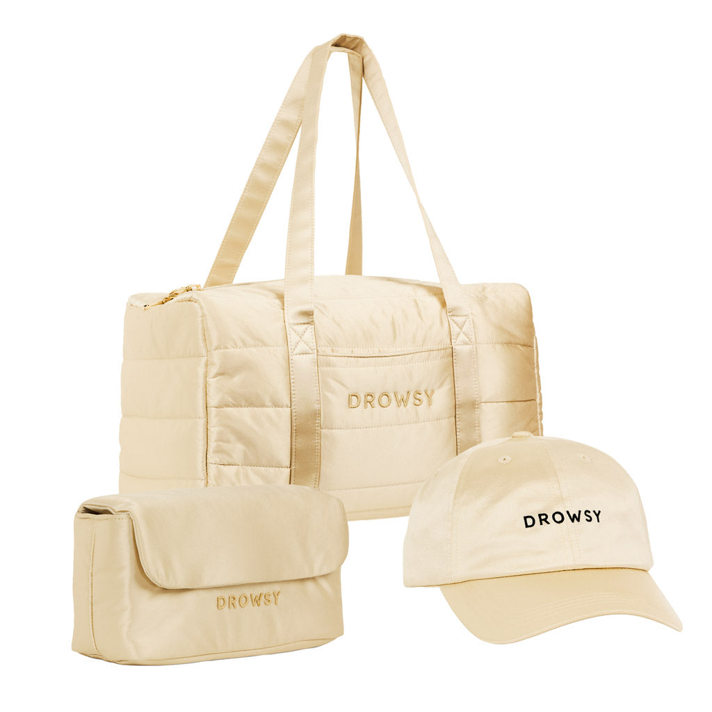 The Drowsy Weekend Collection - Dusty Gold features a duffel bag, toiletry bag, and baseball cap—each in beige with “DROWSY” embroidered in black—neatly arranged on a white background.