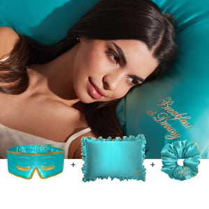 A woman lies on a teal satin Drowsy Hollywood Experience Bundle pillow reading "Breakfast at Draper," with a matching ruffled pillowcase, scrunchie, and gold eyelash-embroidered sleep mask in the same luxurious teal satin.