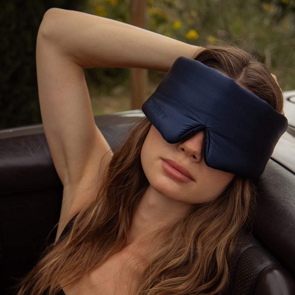 A woman with long brown hair sits in a car seat, wearing the Drowsy Sleepover bundle sleep mask in Midnight Blue. One arm rests behind her head as greenery and flowers appear in the background.