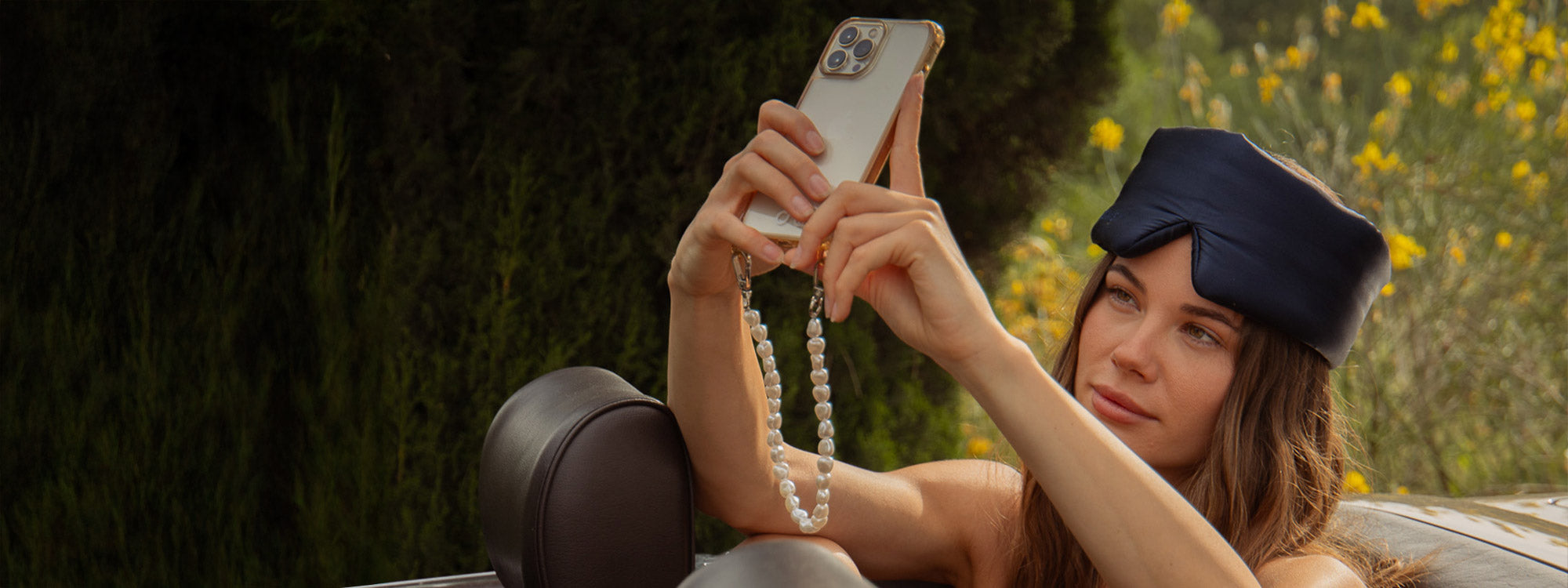 A woman in a car takes a selfie with her smartphone. She is wearing a navy sleep mask pushed up on her head, and there are green trees and yellow flowers in the background.
