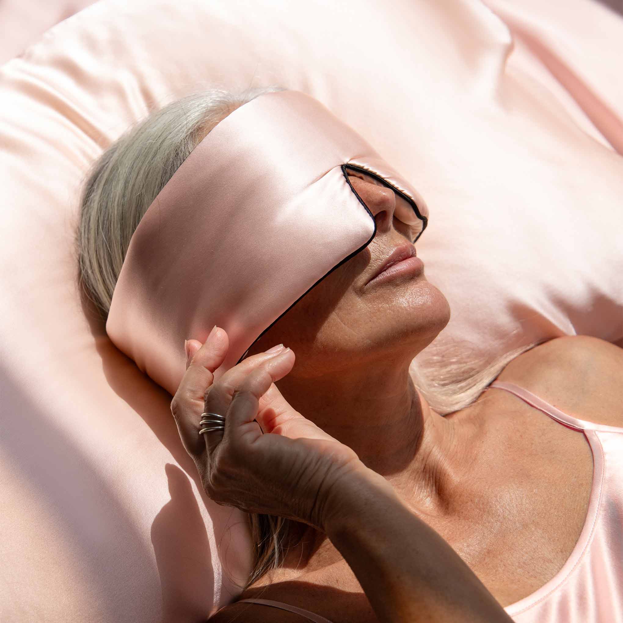Model lying on a pastel pink coloured Drowsy silk pillowcase with a pink silk sleep mask covering her eyes