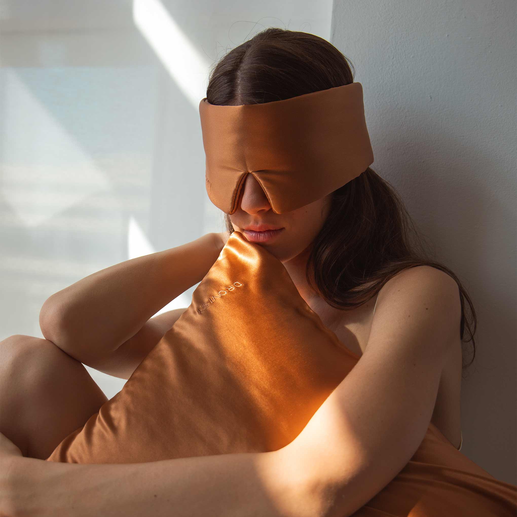 Female model holding terracotta coloured Drowsy silk pillowcase with a Drowsy sleep mask covering her eyes