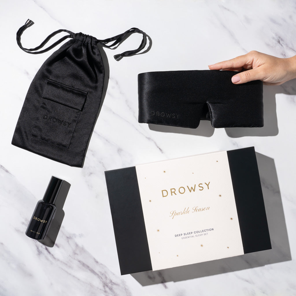 The Black Jade - Deep Sleep Collection by us.drowsysleepco features a luxury silk sleep mask, black silk carry pouch, and deep sleep pillow spray displayed on white marble, with a hand elegantly holding the mask.