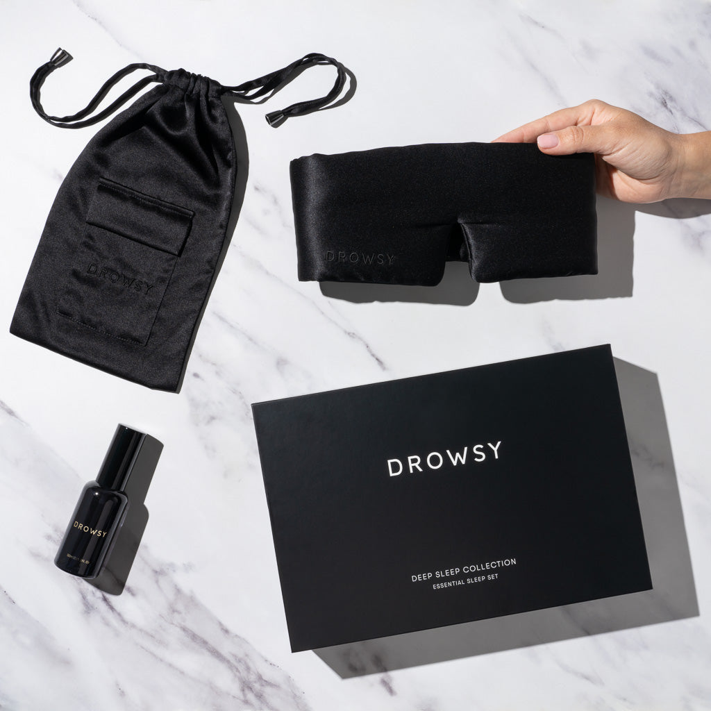 A hand holds a us.drowsysleepco Black Jade - Deep Sleep Collection silk sleep mask beside a black satin pouch, deep sleep pillow spray, and signature box, arranged on a white surface.