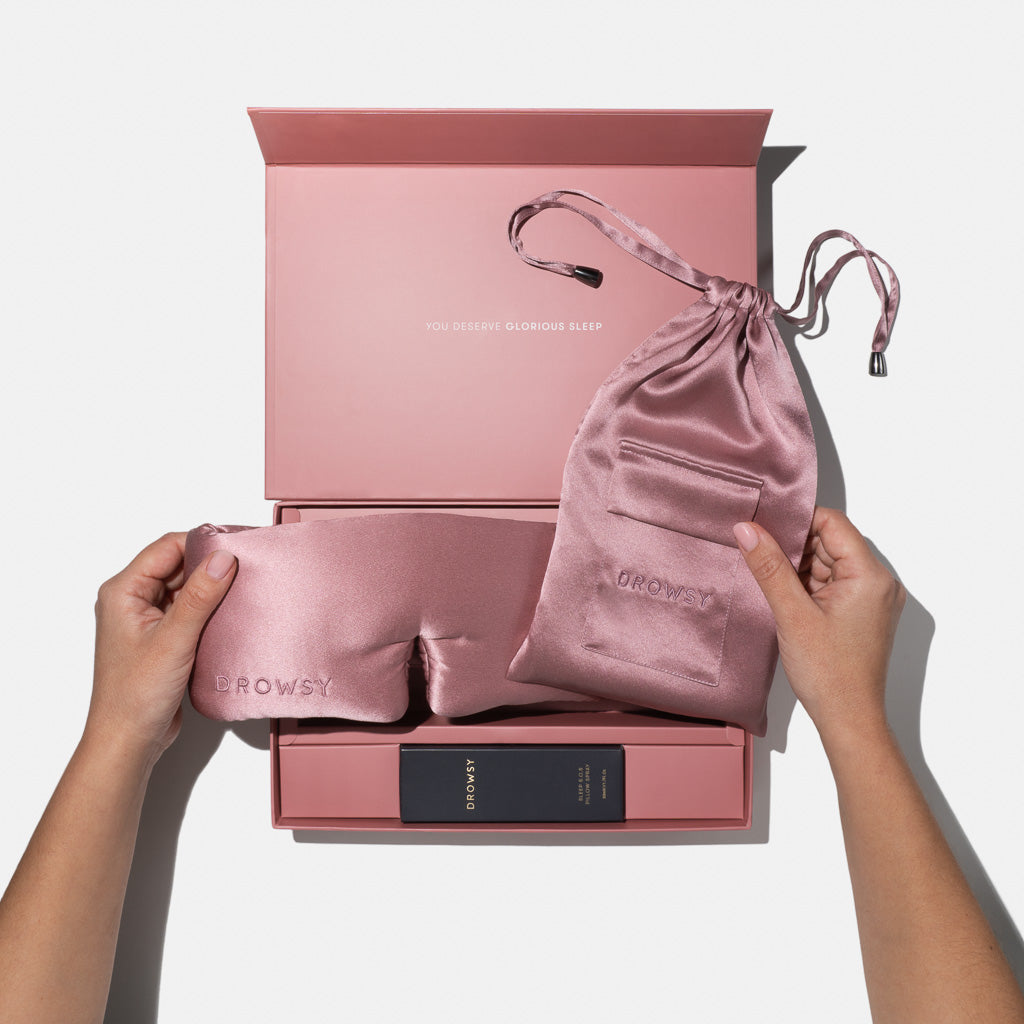 A person holds the us.drowsysleepco Damask Rose - Deep Sleep Collection silk sleep mask and pouch above an open pink gift box with sleep essentials. The box lid says, YOU DESERVE GLORIOUS SLEEP.