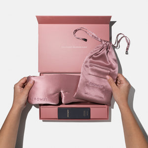 A person holds the us.drowsysleepco Damask Rose - Deep Sleep Collection silk sleep mask and pouch above an open pink gift box with sleep essentials. The box lid says, YOU DESERVE GLORIOUS SLEEP.