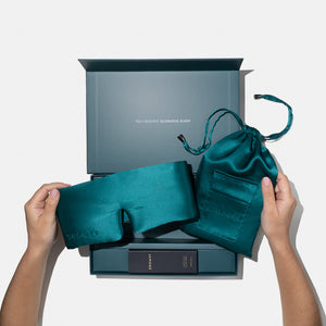 A person holds a Green Sapphire - Deep Sleep Collection silk sleep mask and drawstring pouch by us.drowsysleepco above an open gift box filled with sleep essentials, with "YOU DESERVE GLORIOUS SLEEP" printed inside the lid.