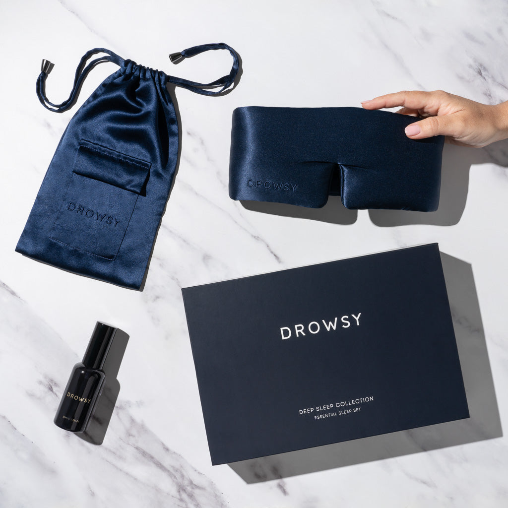 A hand holds a luxury silk sleep mask with a matching blue pouch, black pillow spray, and the us.drowsysleepco Midnight Blue – Deep Sleep Collection box, all displayed on a white marble surface.