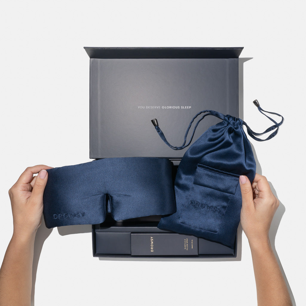 A person holds the us.drowsysleepco Midnight Blue - Deep Sleep Collection silk sleep mask and blue pouch above an open gift box reading 