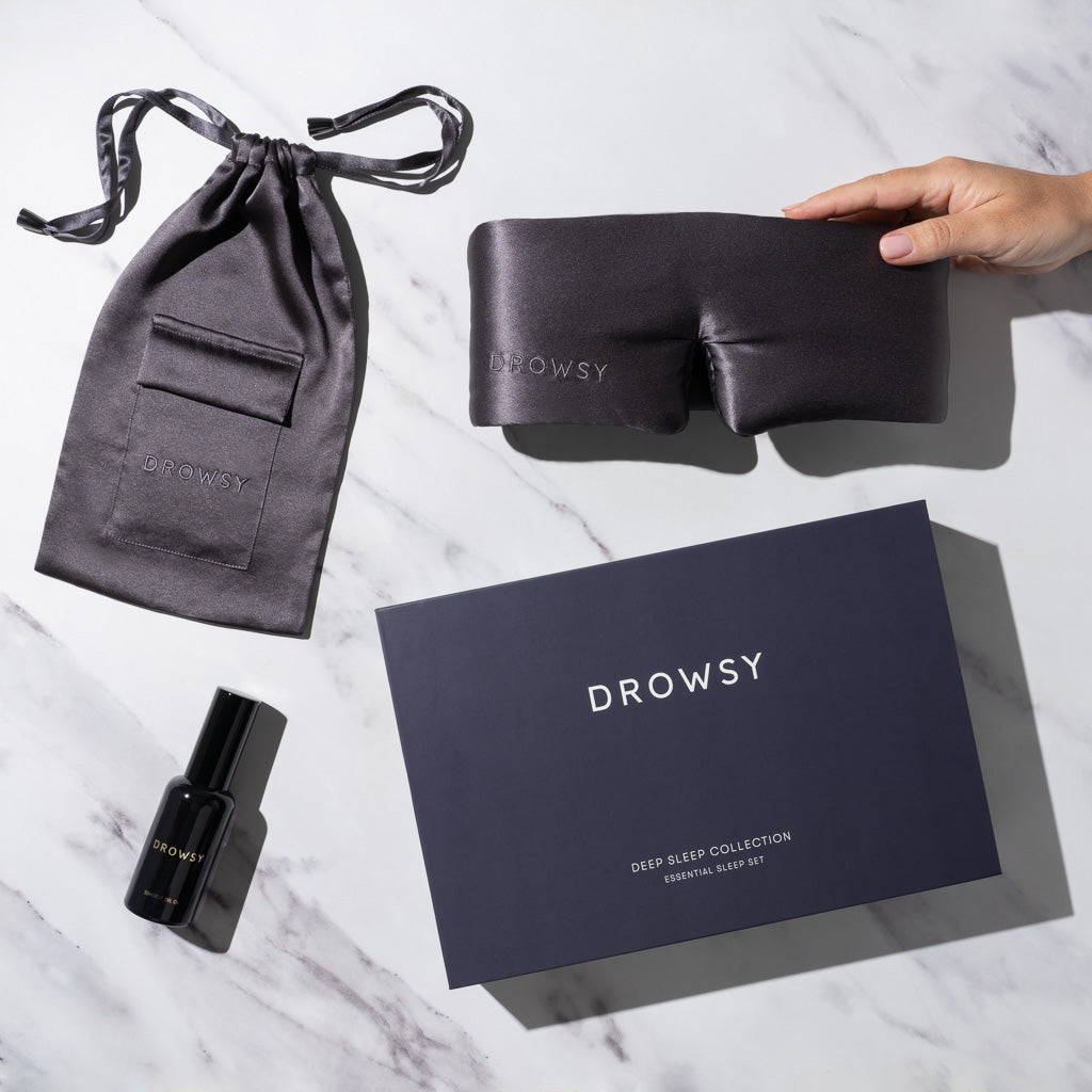 A hand holds a luxury silk sleep mask with a matching pouch, pillow spray, and a dark blue us.drowsysleepco box labeled "Moonlight Shadow - Deep Sleep Collection," all displayed on white marble.