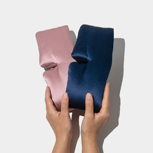 Two hands hold the Drowsy Lovers Pack, featuring a pink and a navy blue luxury silk sleep mask embossed with "DROWSY," against a plain white background.