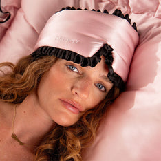 A woman with wavy hair lies on a pink satin pillow, wearing the drowsysleepco Sleep and the City Pink & Black mulberry silk blackout sleep mask with 