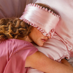 A person with wavy hair lies on a pink satin pillow, wearing drowsysleepco’s Sleep and the City Powder Pink mulberry silk blackout sleep mask, and a pink satin top, appearing relaxed and comfortable.